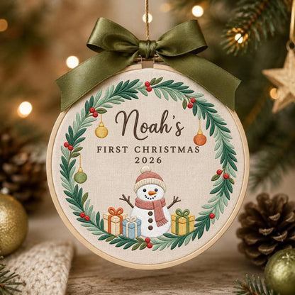 May include: A round embroidery hoop ornament featuring a beige fabric center with a green wreath, red berries, and small hanging ornaments. The text reads Noah's FIRST CHRISTMAS 2026 above a snowman with three gifts. An olive green satin bow tops the hoop.