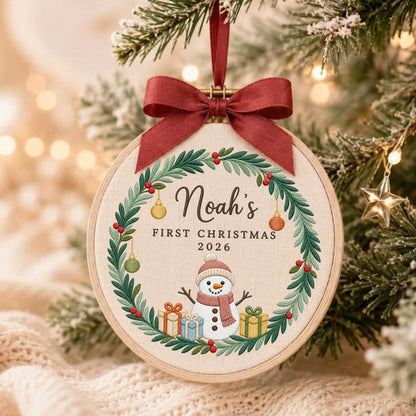 May include: A round wooden embroidery hoop ornament featuring a fabric center with a printed green wreath, a snowman, and gift boxes. The text reads Noah's FIRST CHRISTMAS 2026. A dark red satin ribbon bow is tied at the top for hanging purposes.