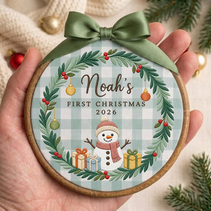 May include: A round wooden embroidery hoop ornament featuring a light green and white buffalo check fabric background. It displays a printed wreath of green pine branches with red berries, a snowman, gifts, and the text Noah's FIRST CHRISTMAS 2026.