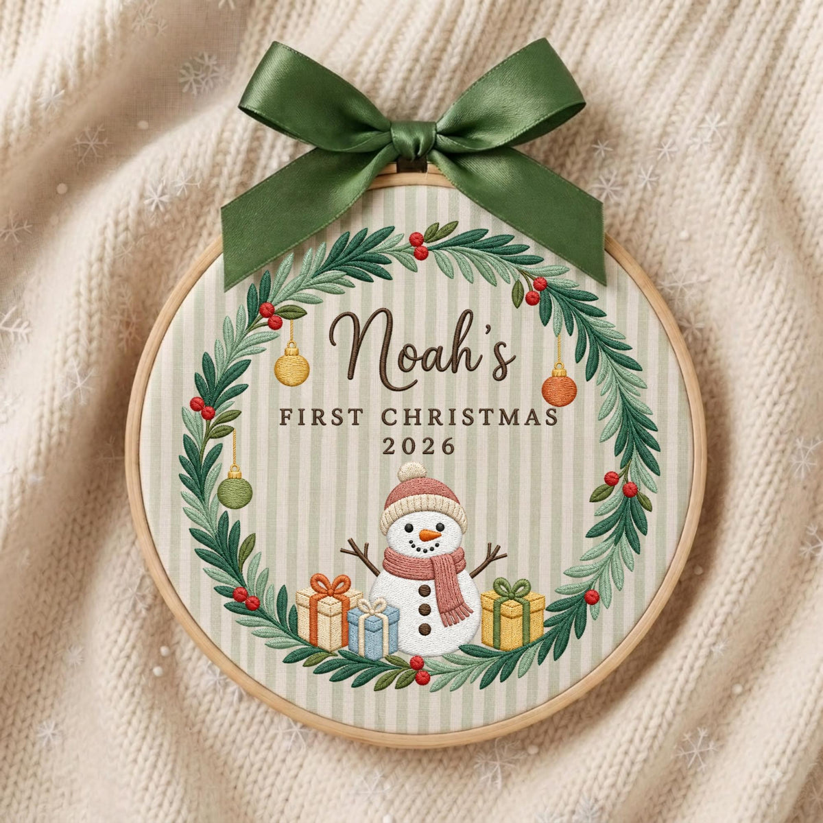 May include: A round wooden embroidery hoop featuring a fabric background with light green stripes. A green satin bow sits at the top. The design includes a wreath of green leaves and red berries, a snowman, gifts, and the text Noah's FIRST CHRISTMAS 2026.
