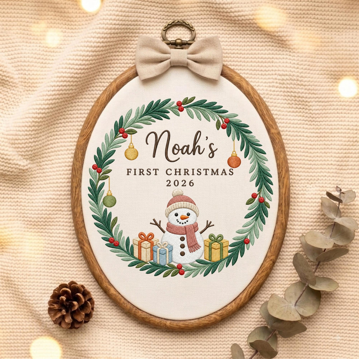 May include: An oval wooden embroidery hoop containing white fabric with a stitched wreath of green leaves and red berries. The text reads Noah's FIRST CHRISTMAS 2026 above a snowman with a pink hat and scarf, surrounded by three colorful gift boxes.