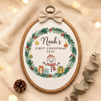 May include: An oval wooden embroidery hoop containing white fabric with a stitched wreath of green leaves and red berries. The text reads Noah's FIRST CHRISTMAS 2026 above a snowman with a pink hat and scarf, surrounded by three colorful gift boxes.