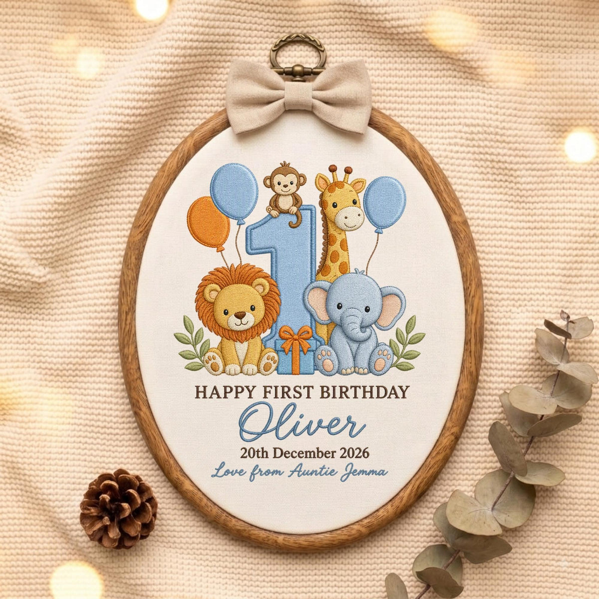 May include: An oval wooden embroidery hoop containing white fabric with a colorful embroidered design of a lion, giraffe, elephant, and monkey around a large blue number one. Text reads: HAPPY FIRST BIRTHDAY Oliver 20th December 2026 Love from Auntie Jemma.