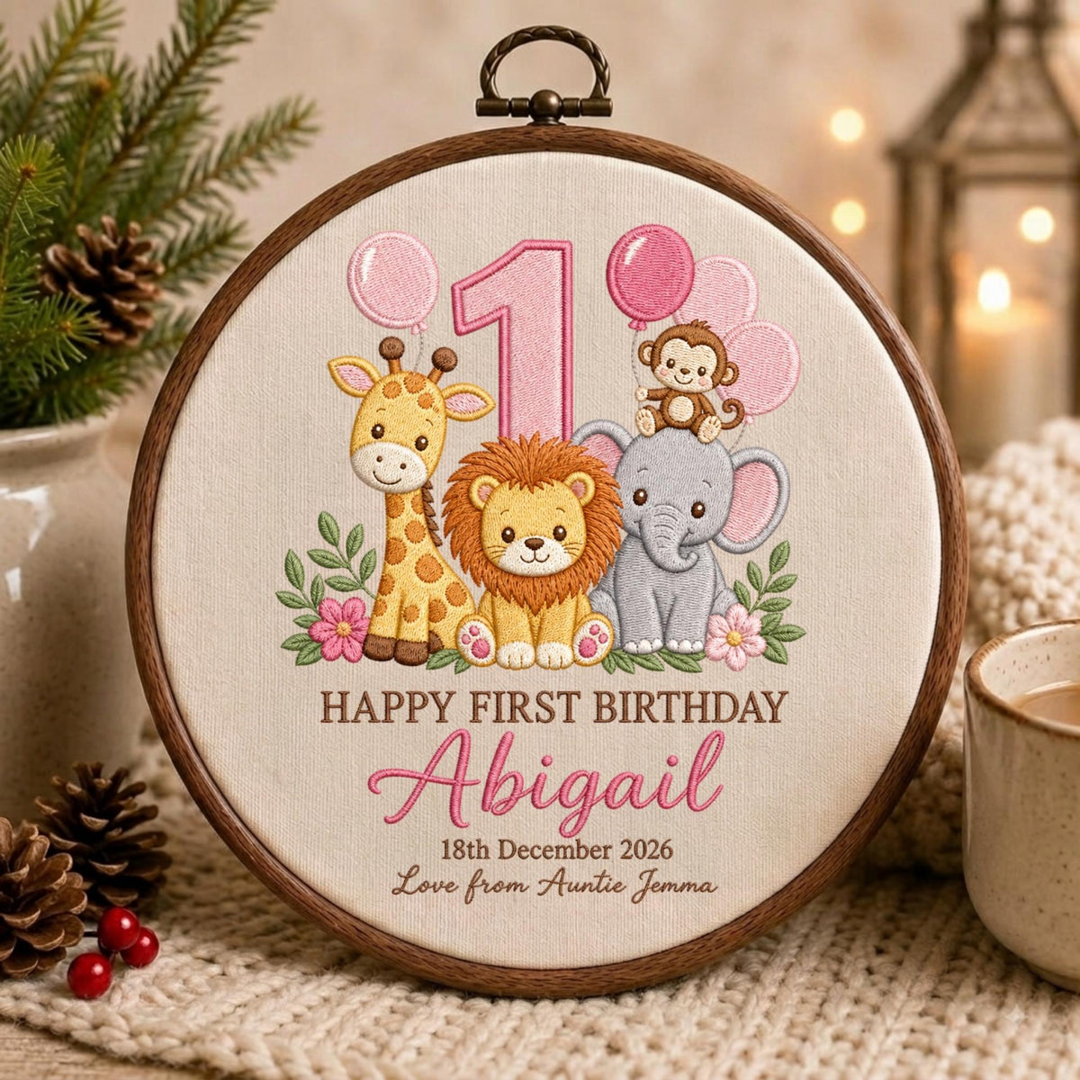 May include: A circular wooden embroidery hoop containing fabric with a stitched design of a giraffe, lion, elephant, and monkey. The design features a large pink number 1, pink balloons, and the text Happy First Birthday Abigail 18th December 2026 Love from Auntie Jemma.