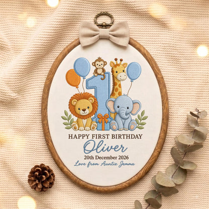 May include: An oval embroidery hoop featuring a light blue number one, a lion, a giraffe, an elephant, and a monkey. The text reads HAPPY FIRST BIRTHDAY, Oliver, 20th December 2026, and Love from Auntie Jemma. A beige bow is attached to the top.