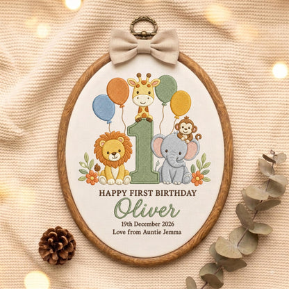May include: An oval embroidery hoop featuring a white fabric center with a stitched design of a giraffe, lion, monkey, and elephant around a large number one. Text reads HAPPY FIRST BIRTHDAY, Oliver, 19th December 2026, Love from Auntie Jemma.