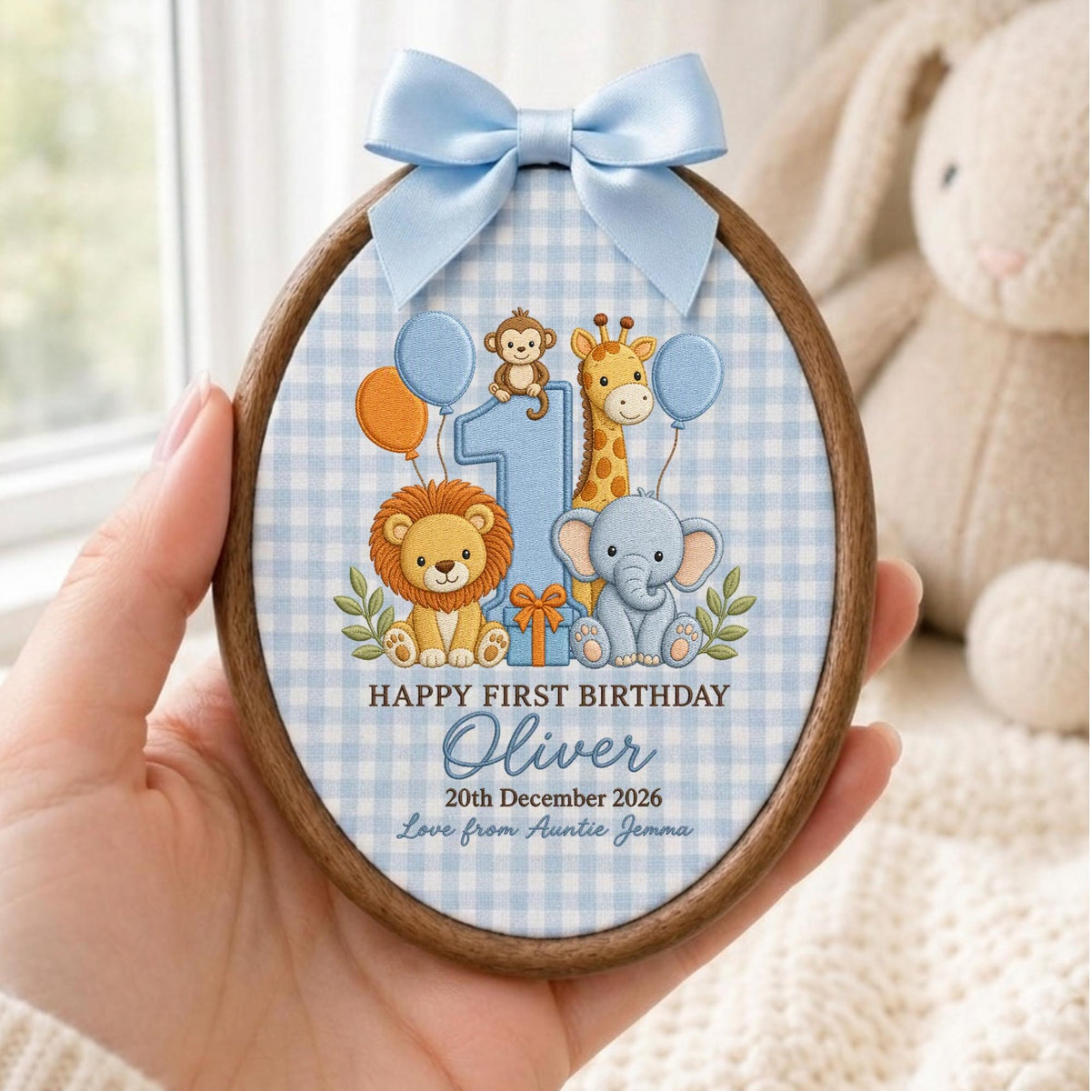 May include: An oval wooden embroidery hoop featuring a light blue gingham fabric background. The design includes a large number one, a lion, a giraffe, an elephant, a monkey, and balloons. Text reads: HAPPY FIRST BIRTHDAY Oliver 20th December 2026 Love from Auntie Jemma. A light blue satin bow is attached at the top.