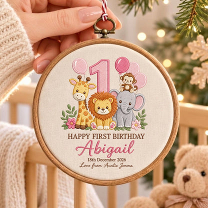 May include: A round wooden embroidery hoop featuring a fabric insert with a printed design of a giraffe, lion, and elephant. The text reads 1, HAPPY FIRST BIRTHDAY, Abigail, 18th December 2026, and Love from Auntie Jemma, with pink balloons.