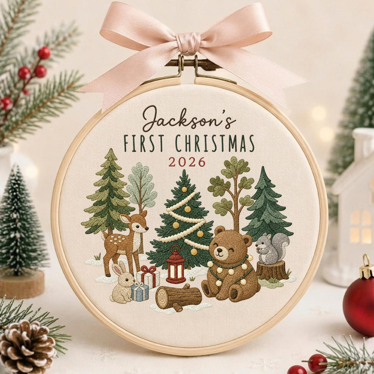 May include: A round wooden embroidery hoop featuring a cream fabric background with the text Jackson's FIRST CHRISTMAS 2026. The design includes embroidered forest animals, trees, and gifts, topped with a light pink satin ribbon bow.