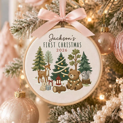 May include: A round wooden embroidery hoop ornament featuring a light pink satin ribbon bow at the top. The fabric center displays embroidered forest animals including a deer, rabbit, bear, and squirrel, with trees, gifts, and the text Jackson's FIRST CHRISTMAS 2026.