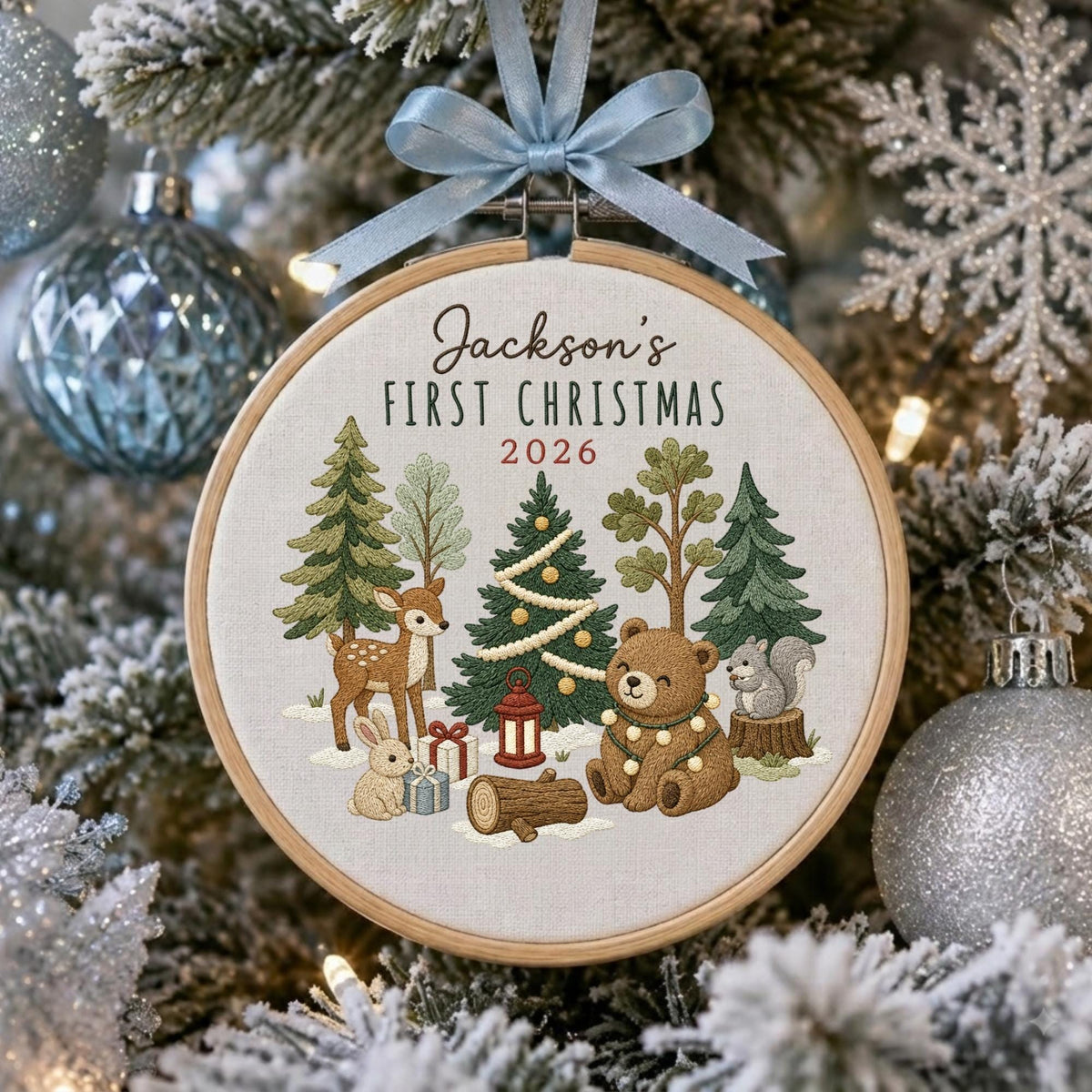 May include: A round wooden embroidery hoop ornament featuring a light fabric background with the text Jackson's FIRST CHRISTMAS 2026. The design includes embroidered forest animals, trees, a decorated pine tree, and gifts, topped with a light blue ribbon.