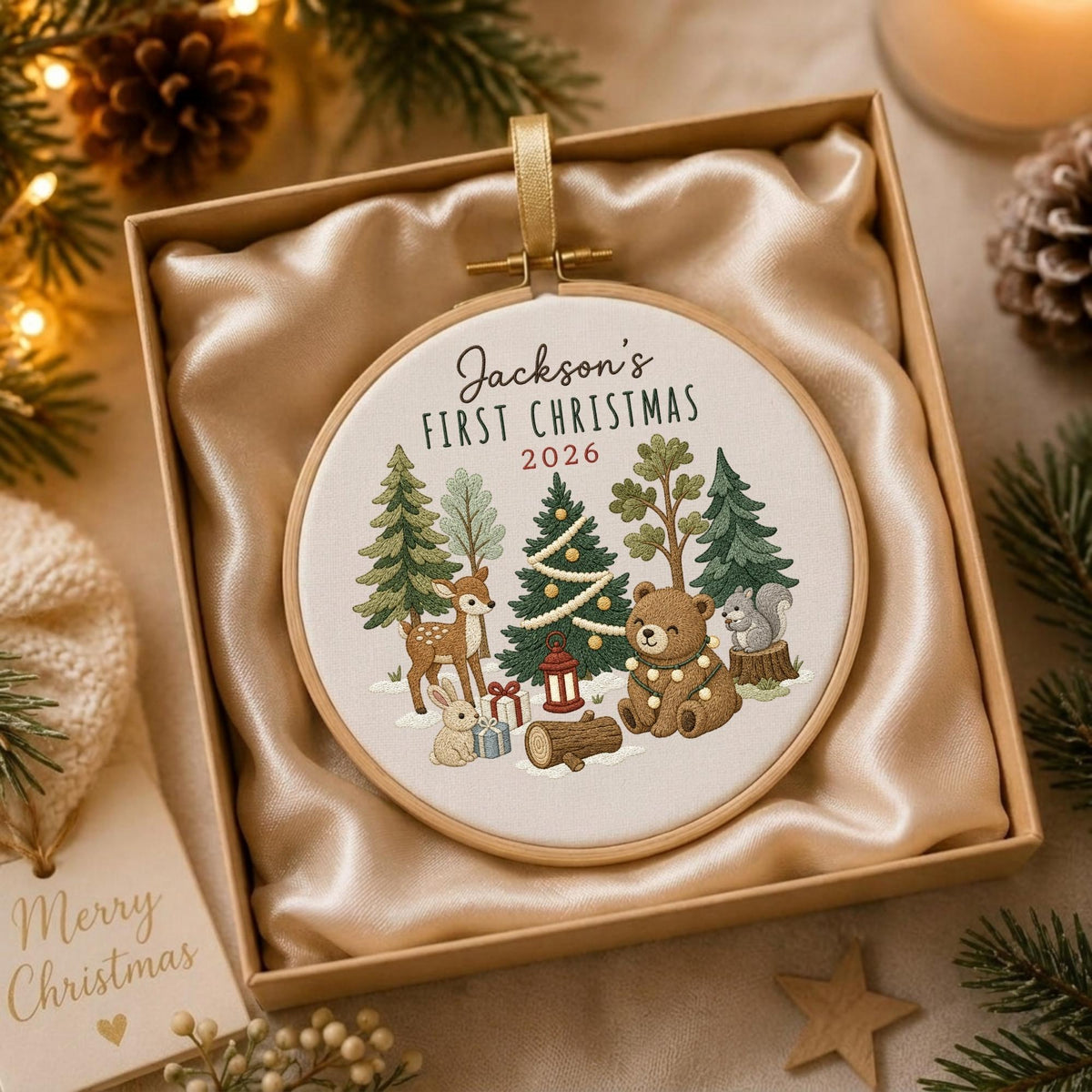 May include: A round wooden embroidery hoop ornament featuring a cream fabric center with the embroidered text Jackson's FIRST CHRISTMAS 2026. The design includes a central Christmas tree, a brown bear, a deer, a rabbit, and a squirrel.