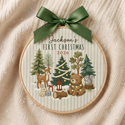 May include: A circular wooden embroidery hoop featuring a light green and white striped fabric background. The text reads Jackson's FIRST CHRISTMAS 2026. Embroidered forest animals, trees, gifts, and a lantern are displayed, topped with a dark green satin bow.