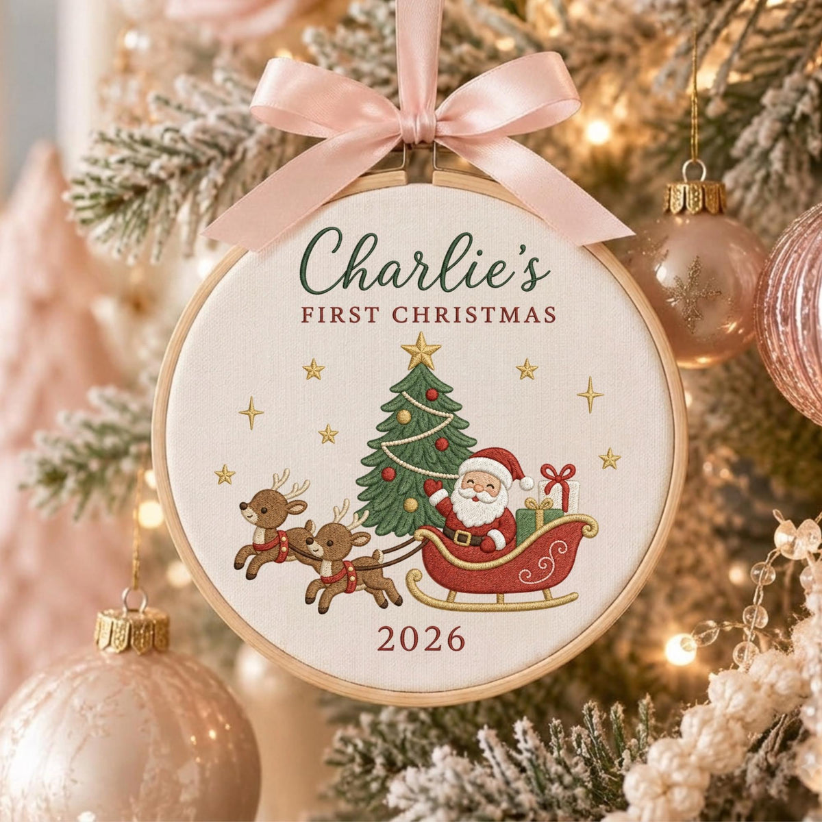May include: A round wooden embroidery hoop ornament featuring a light pink satin ribbon bow at the top. The fabric center displays embroidered text reading Charlie's FIRST CHRISTMAS and 2026, with a green Christmas tree, Santa in a red sleigh, and two reindeer.