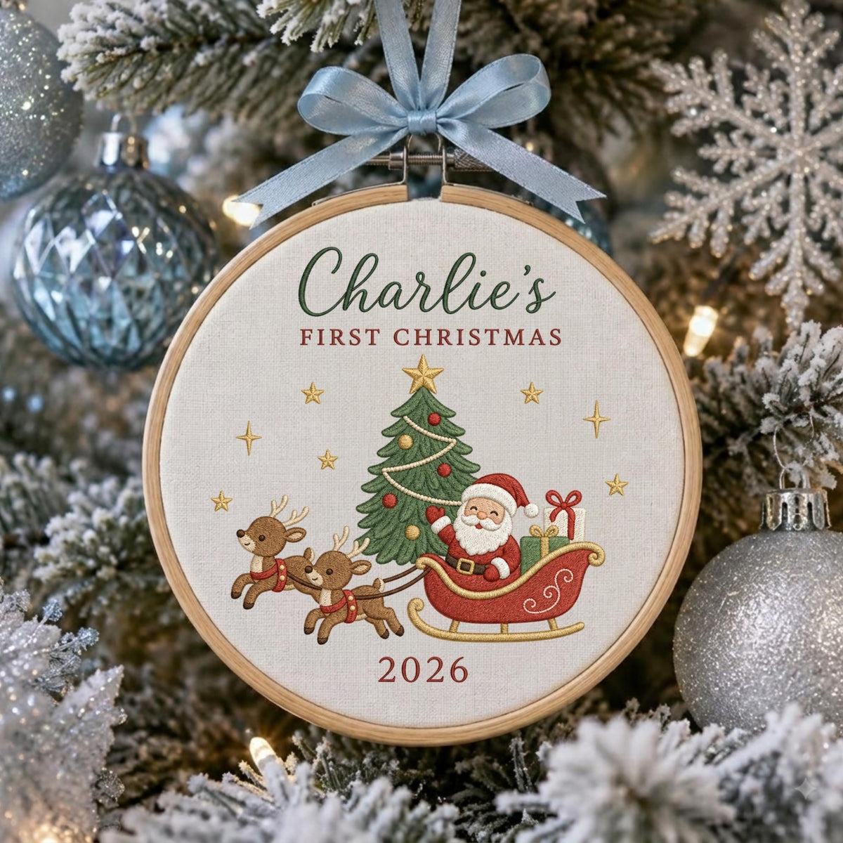 May include: A round wooden embroidery hoop ornament featuring a light fabric center with embroidered text Charlie's FIRST CHRISTMAS and 2026. The design includes a green Christmas tree, Santa Claus in a red sleigh, and two reindeer, topped with a light blue ribbon.