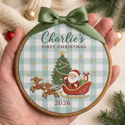 May include: A round wooden embroidery hoop ornament featuring a light blue and white gingham fabric background. The design includes a green Christmas tree, Santa Claus in a red sleigh pulled by two reindeer, and the text Charlie's FIRST CHRISTMAS 2026. A sage green satin ribbon bow is tied at the top.