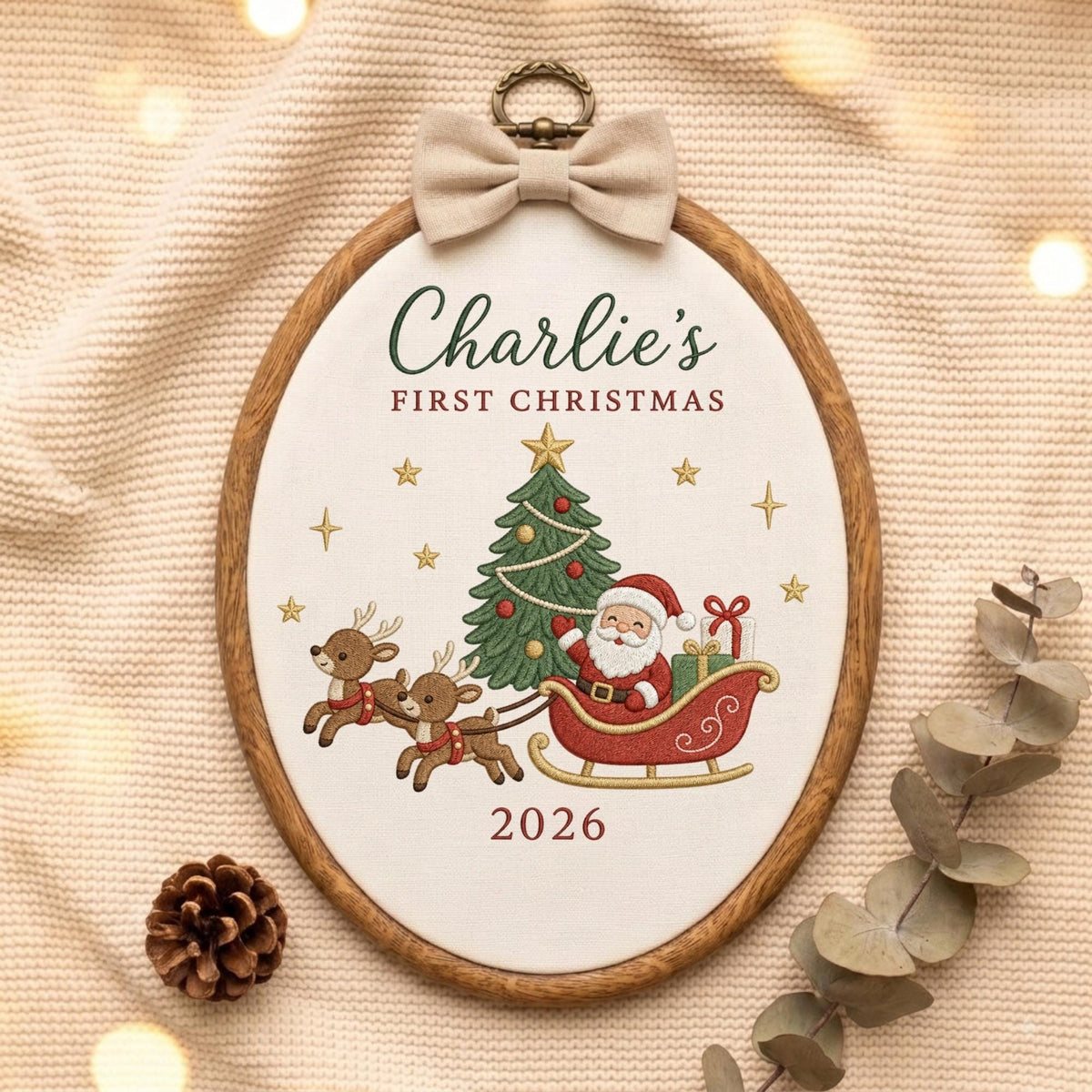 May include: An oval embroidery hoop featuring a white fabric center with the embroidered text Charlie's FIRST CHRISTMAS and 2026. The design includes a green Christmas tree, Santa Claus in a red sleigh, two reindeer, and gold stars. A beige bow sits at the top.