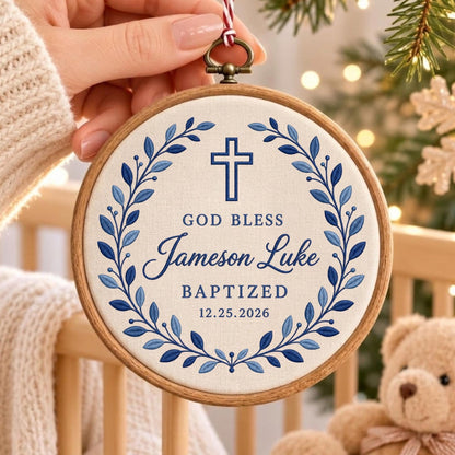 May include: A circular wooden embroidery hoop containing white fabric with blue embroidered text reading GOD BLESS Jameson Luke BAPTIZED 12.25.2026. A blue cross sits above the text, surrounded by a blue leaf wreath. A red and white string hangs from the top.