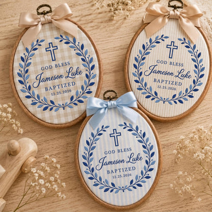 May include: Three oval wooden embroidery hoops featuring fabric inserts with blue embroidered wreaths, crosses, and the text "GOD BLESS Jameson Luke BAPTIZED 12.25.2026". Each hoop is topped with a decorative satin ribbon bow in beige or light blue.
