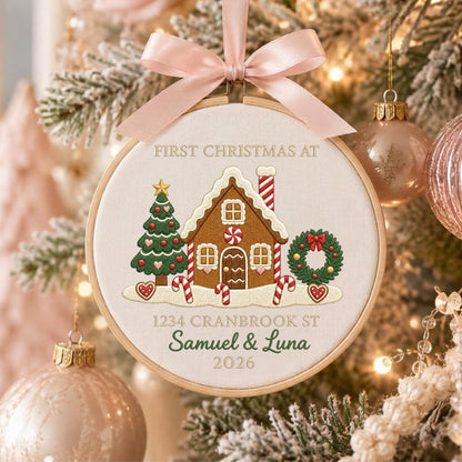 May include: A round embroidery hoop ornament featuring a gingerbread house, a Christmas tree, and a wreath. The text reads FIRST CHRISTMAS AT, 1234 CRANBROOK ST, Samuel &amp; Luna, and 2026. A light pink satin ribbon bow is tied at the top of the hoop.