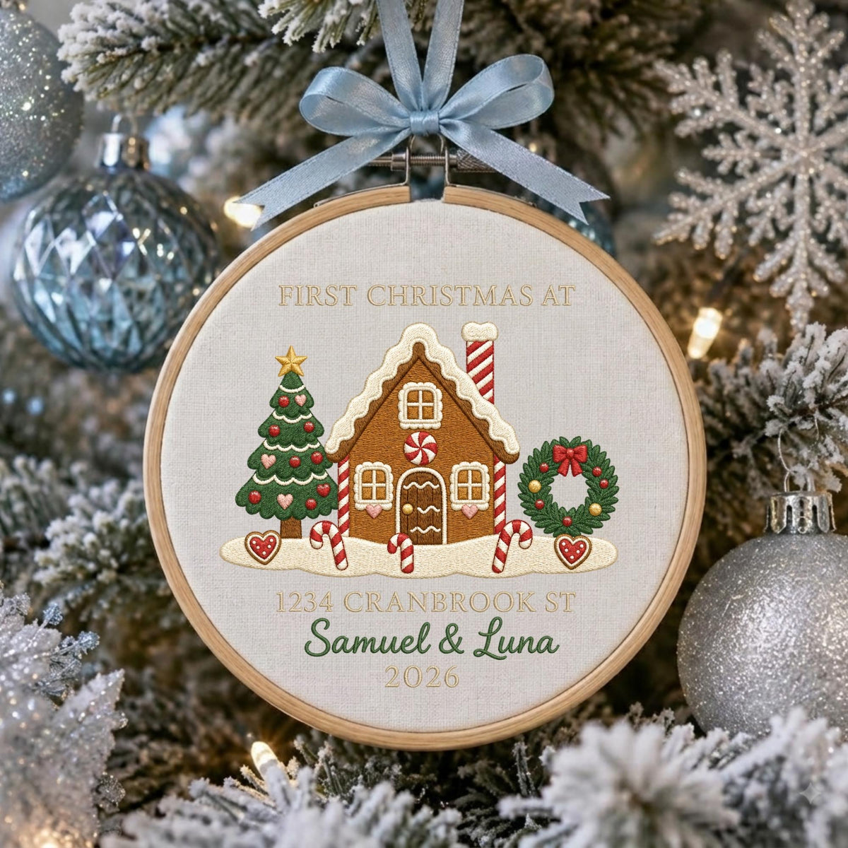May include: A round wooden embroidery hoop ornament featuring a light fabric background with an embroidered gingerbread house, a Christmas tree, and a wreath. The text reads FIRST CHRISTMAS AT 1234 CRANBROOK ST, Samuel &amp; Luna, and 2026.