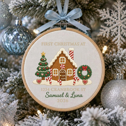 May include: A round wooden embroidery hoop ornament featuring a light fabric background with an embroidered gingerbread house, a Christmas tree, and a wreath. The text reads FIRST CHRISTMAS AT 1234 CRANBROOK ST, Samuel &amp; Luna, and 2026.