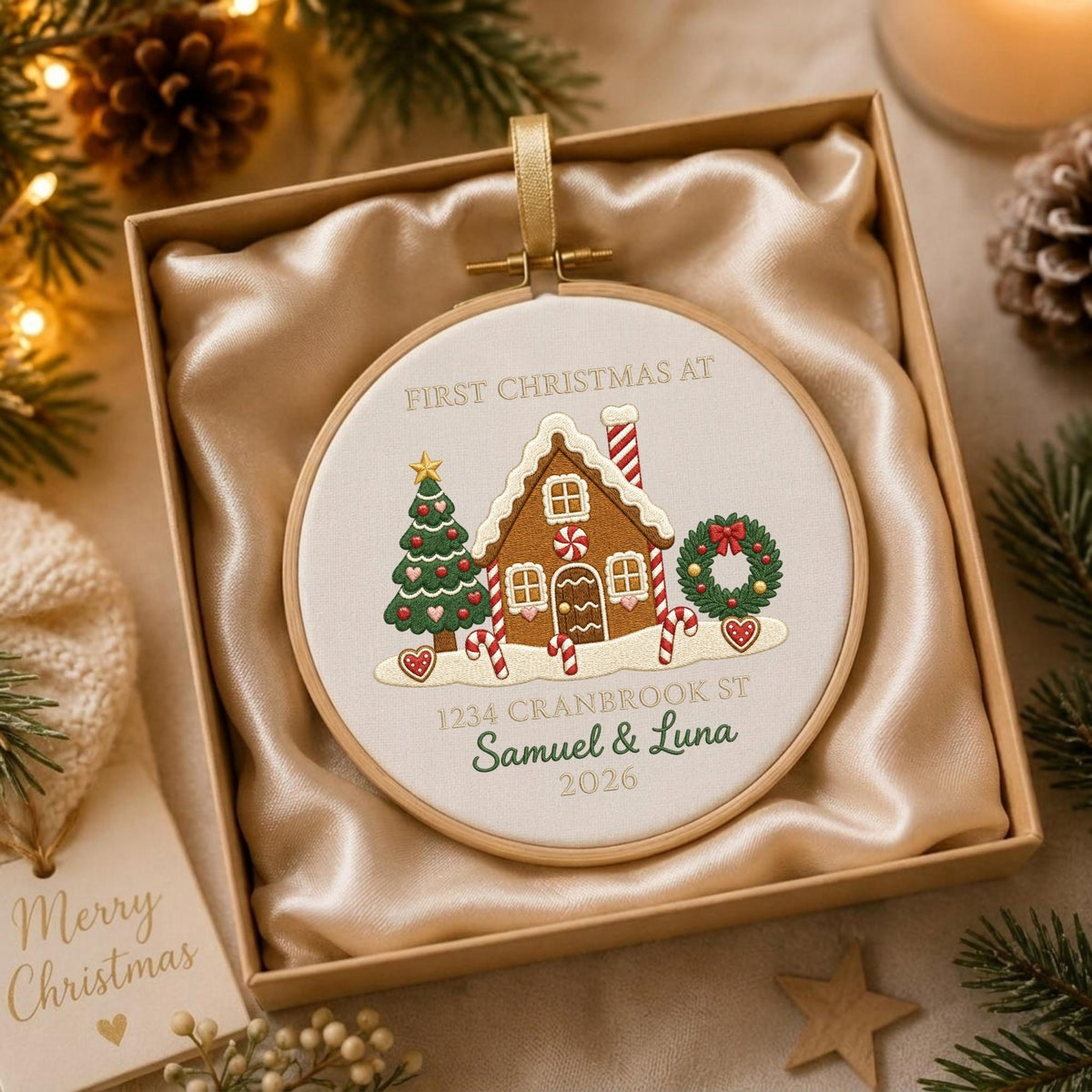 May include: A round wooden embroidery hoop ornament featuring a gingerbread house, a Christmas tree, and a wreath. The text reads FIRST CHRISTMAS AT 1234 CRANBROOK ST SAMUEL &amp; LUNA 2026. The ornament is presented in a square box with champagne satin fabric.
