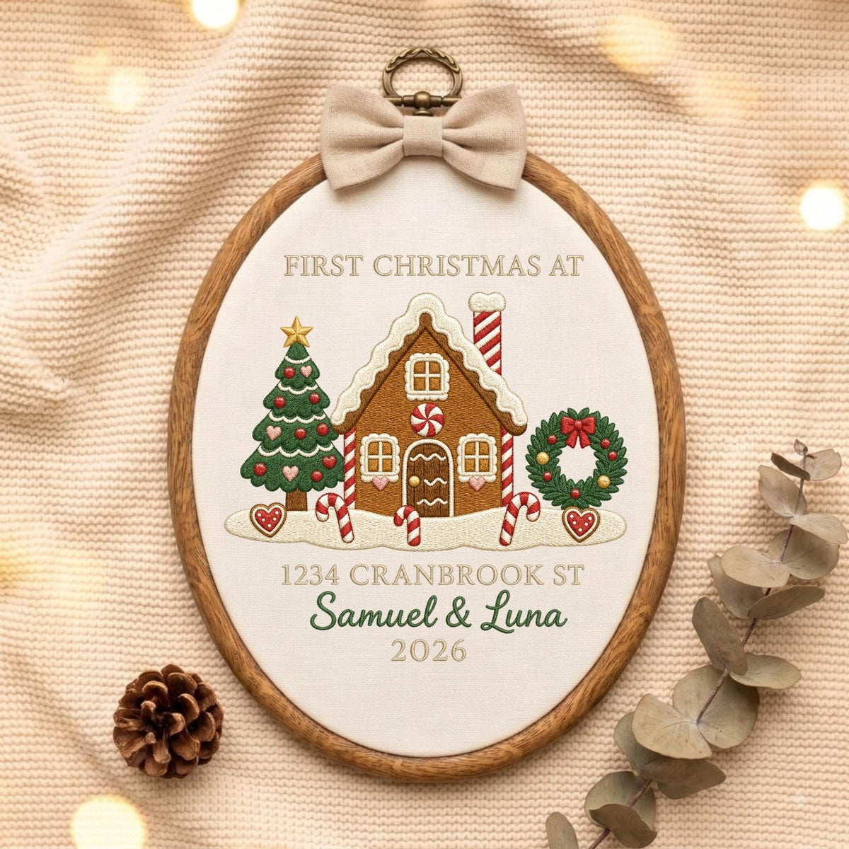 May include: An oval wooden embroidery hoop containing white fabric with a gingerbread house, Christmas tree, and wreath design. Text reads: FIRST CHRISTMAS AT, 1234 CRANBROOK ST, Samuel &amp; Luna, 2026. A beige fabric bow is attached to the top.