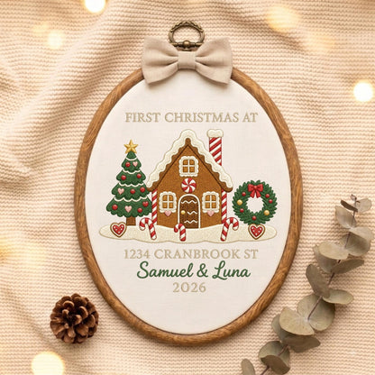 May include: An oval wooden embroidery hoop containing white fabric with a gingerbread house, Christmas tree, and wreath design. Text reads: FIRST CHRISTMAS AT, 1234 CRANBROOK ST, Samuel &amp; Luna, 2026. A beige fabric bow is attached to the top.