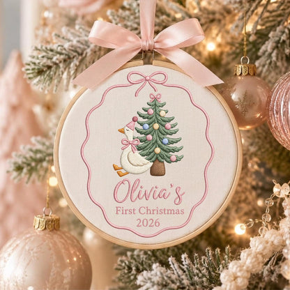 May include: A round wooden embroidery hoop ornament featuring a white fabric center with a pink embroidered border. The design includes a white goose wearing a pink hat and bow, a green Christmas tree with colorful ornaments, and the text Olivia's First Christmas 2026. A light pink satin ribbon bow is attached to the top.