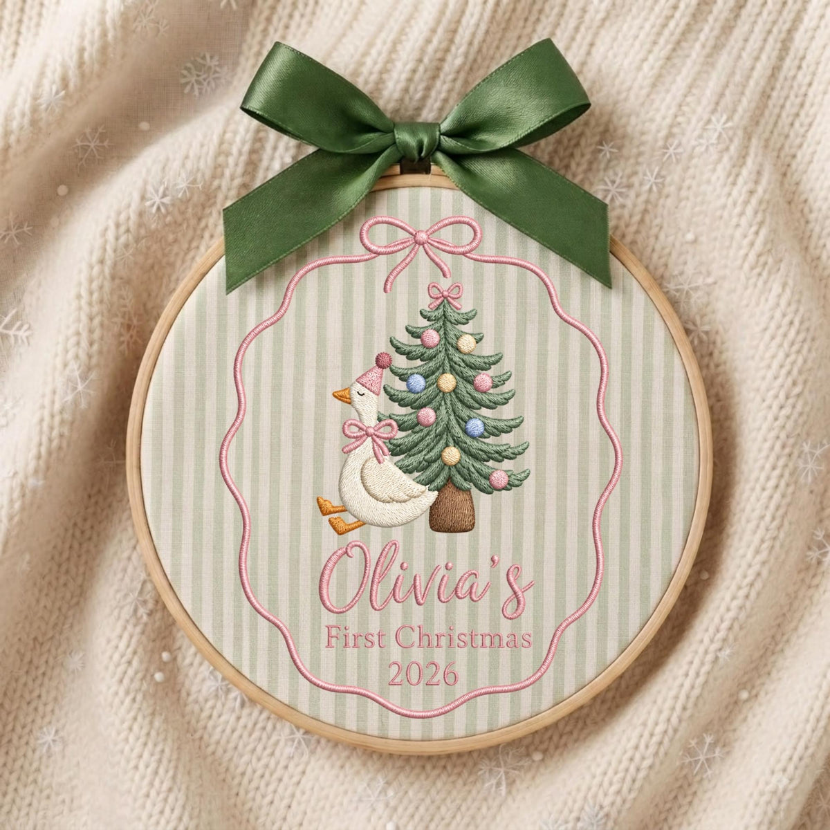 May include: A circular wooden embroidery hoop containing fabric with a light green and white striped pattern. The embroidery features a white goose wearing a pink hat and bow, a decorated Christmas tree, and the text Olivia's First Christmas 2026.