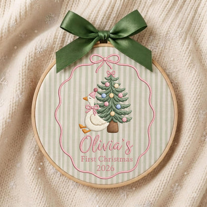 May include: A circular wooden embroidery hoop containing fabric with a light green and white striped pattern. The embroidery features a white goose wearing a pink hat and bow, a decorated Christmas tree, and the text Olivia's First Christmas 2026.