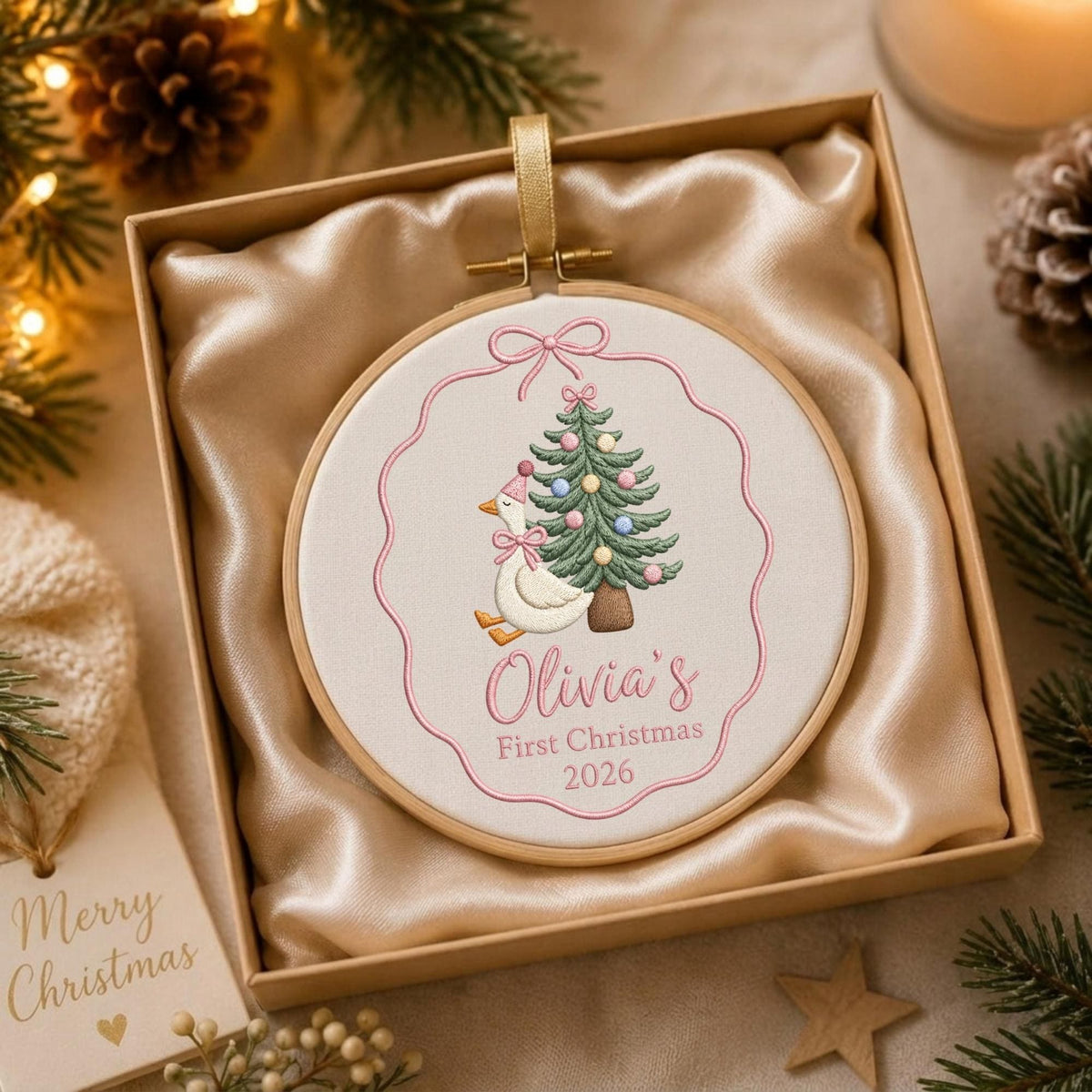 May include: A round wooden embroidery hoop ornament featuring a white fabric center with an embroidered goose wearing a pink hat and bow next to a decorated Christmas tree. The text reads Olivia's First Christmas 2026 in pink script and print.