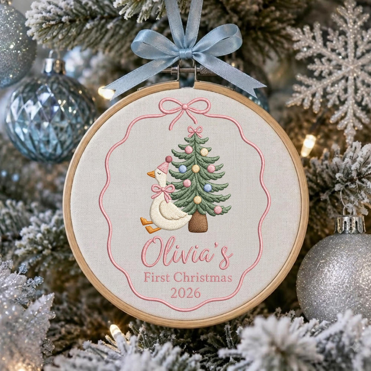 May include: A round wooden embroidery hoop ornament featuring a white fabric background with a pink embroidered border. The design includes a white goose wearing a pink party hat and bow, a decorated Christmas tree, and the text Olivia's First Christmas 2026. A light blue ribbon bow is attached to the top.