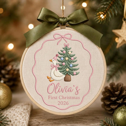 May include: A circular embroidery hoop ornament featuring a white goose wearing a pink party hat and bow, standing next to a decorated Christmas tree. The text reads Olivia's First Christmas 2026. It is topped with a dark olive green satin ribbon bow.