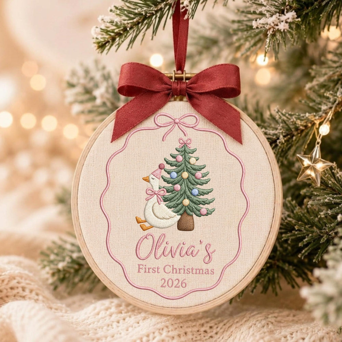 May include: A round wooden embroidery hoop ornament featuring a white fabric center with an embroidered goose wearing a pink party hat and bow, next to a decorated Christmas tree. The text reads Olivia's First Christmas 2026, topped with a dark red ribbon.