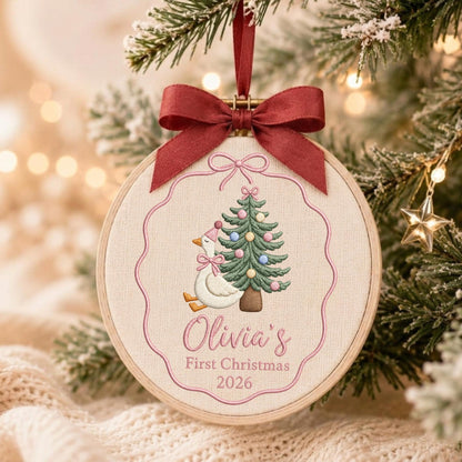May include: A round wooden embroidery hoop ornament featuring a white fabric center with an embroidered goose wearing a pink party hat and bow, next to a decorated Christmas tree. The text reads Olivia's First Christmas 2026, topped with a dark red ribbon.