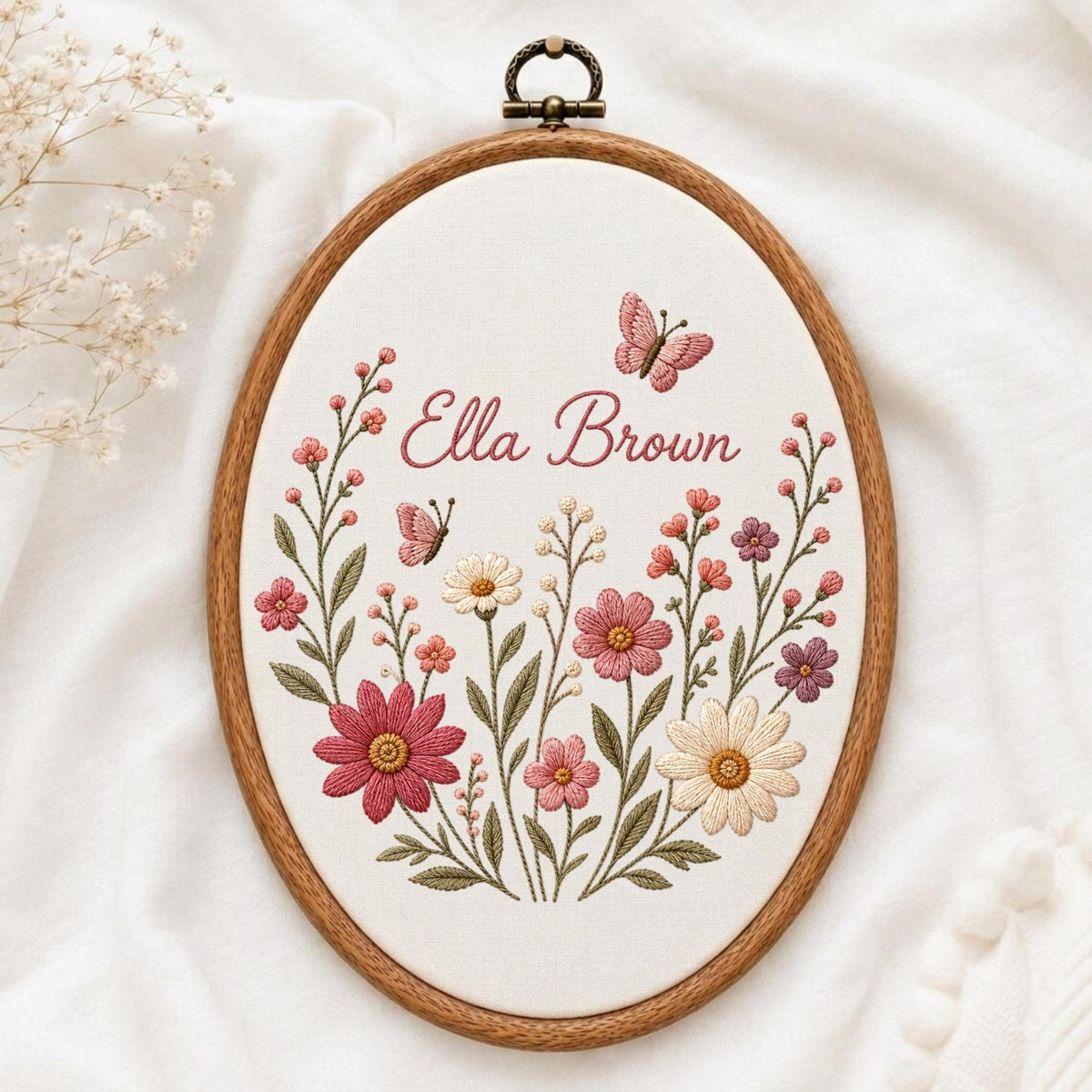 May include: An oval wooden embroidery hoop containing white fabric with hand-stitched floral designs in shades of pink, cream, and green. The embroidery features the name Ella Brown in cursive script, accompanied by two small butterflies.