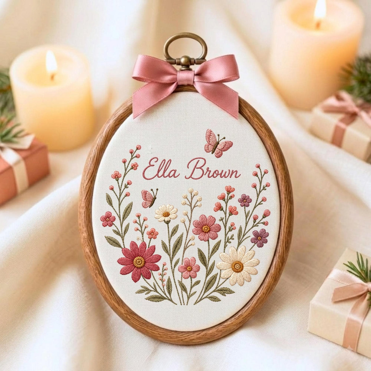 May include: An oval wooden embroidery hoop containing white fabric with floral embroidery in shades of pink, red, and cream. The text "Ella Brown" is stitched in pink script above the flowers, with two small butterflies. A pink satin bow is attached.
