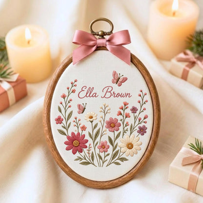 May include: An oval wooden embroidery hoop containing white fabric with floral embroidery in shades of pink, red, and cream. The text "Ella Brown" is stitched in pink script above the flowers, with two small butterflies. A pink satin bow is attached.