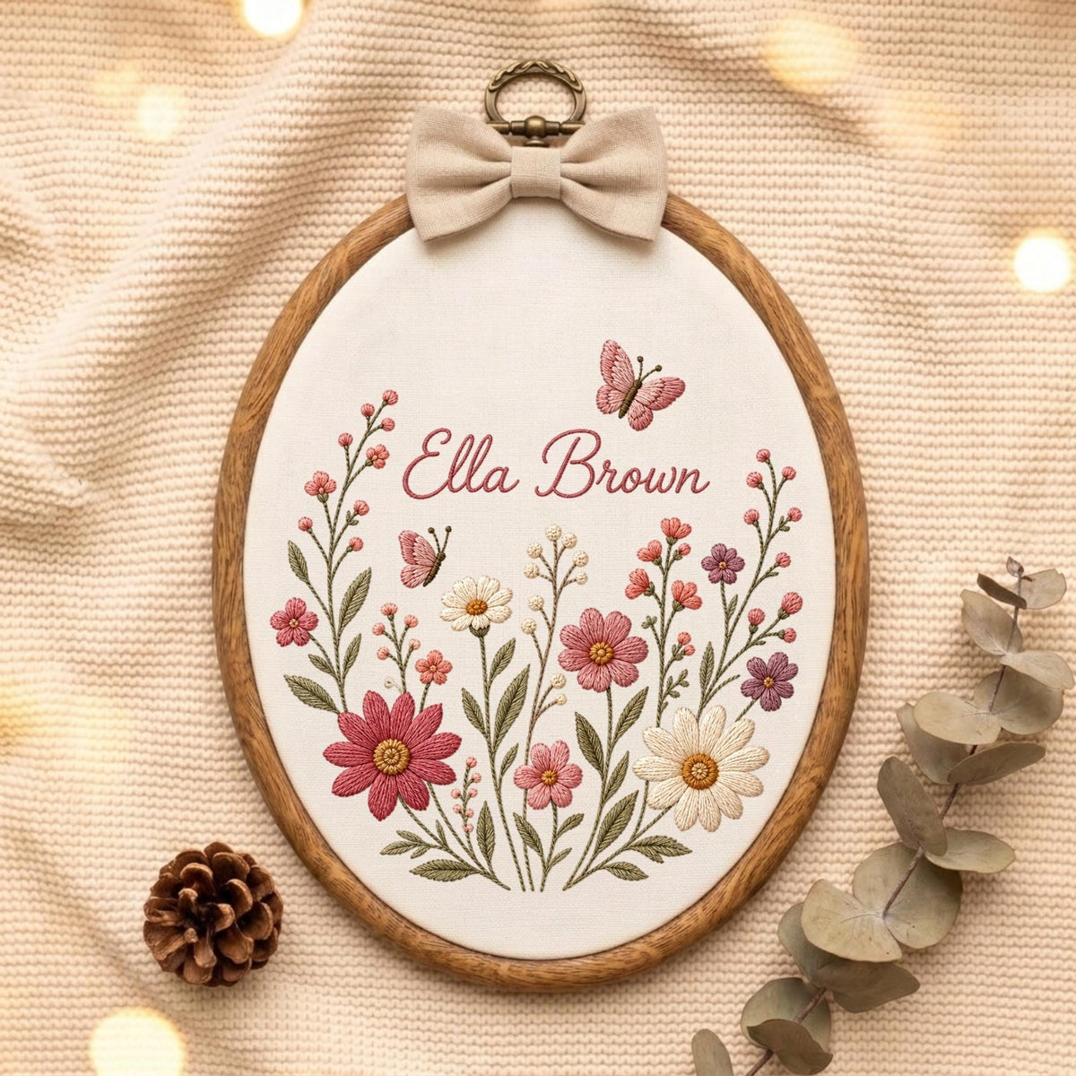 May include: An oval wooden embroidery hoop containing white fabric with a floral design featuring pink, red, and white flowers, green stems, and two butterflies. The name Ella Brown is embroidered in pink script. A beige fabric bow sits at the top.