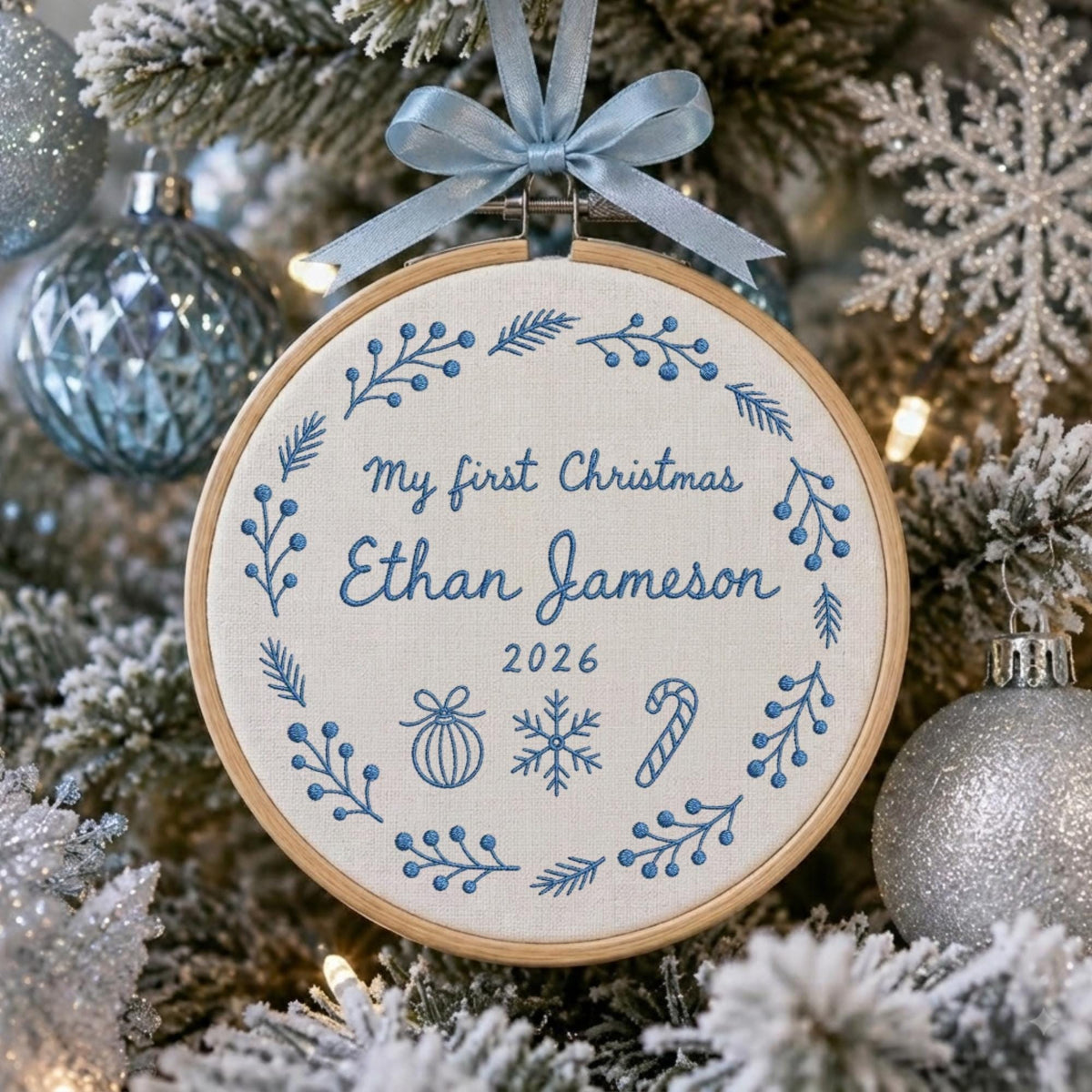 May include: A round wooden embroidery hoop ornament featuring white fabric with blue embroidered text reading My first Christmas Ethan Jameson 2026. The design includes a blue floral wreath, a bauble, a snowflake, and a candy cane, topped with a light blue ribbon.
