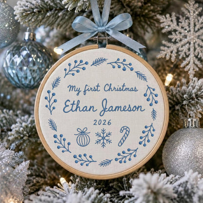 May include: A round wooden embroidery hoop ornament featuring white fabric with blue embroidered text reading My first Christmas Ethan Jameson 2026. The design includes a blue floral wreath, a bauble, a snowflake, and a candy cane, topped with a light blue ribbon.
