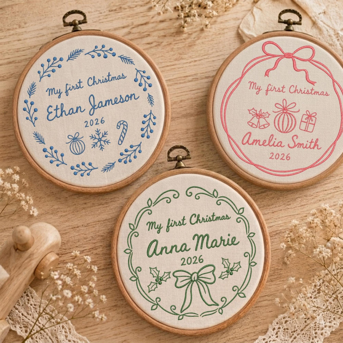 May include: Three circular wooden embroidery hoops containing white fabric with personalized Christmas designs. Each hoop features the text My first Christmas, a name, and the year 2026, embroidered in blue, pink, or green thread with festive motifs.