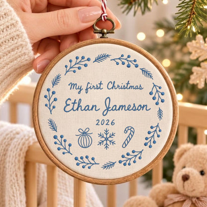 May include: A round wooden embroidery hoop containing off-white fabric with blue embroidered text reading My first Christmas Ethan Jameson 2026. The design features a circular wreath of blue sprigs, a Christmas ornament, a snowflake, and a candy cane.