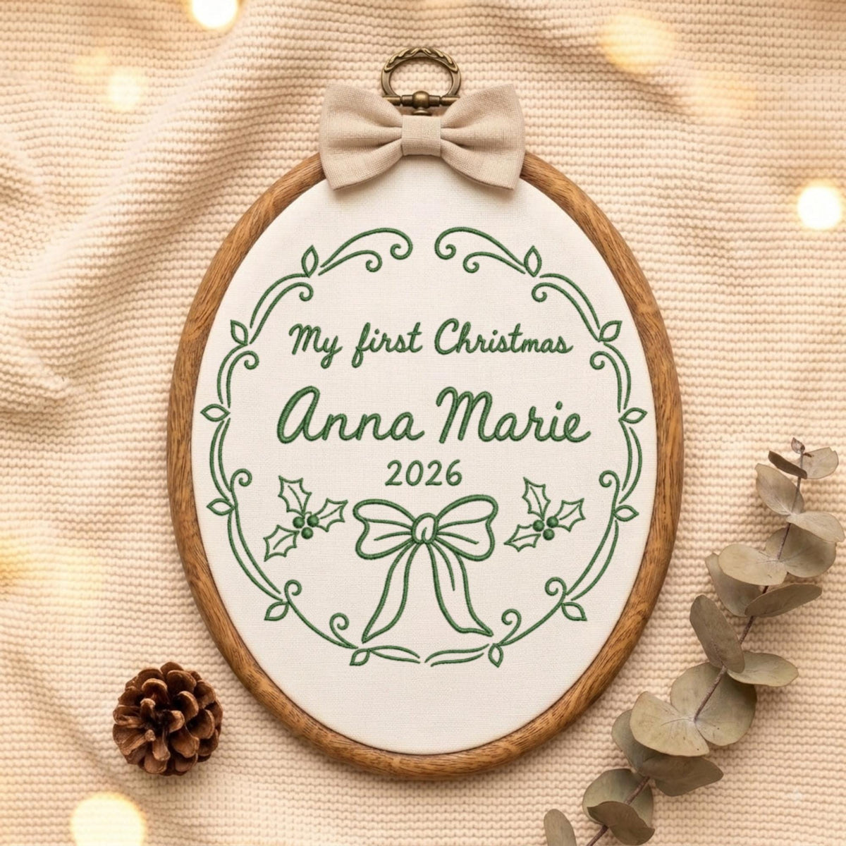 May include: An oval embroidery hoop featuring white fabric with green stitched text reading My first Christmas Anna Marie 2026. The design includes a green floral border, holly leaves, and a bow. A beige fabric bow is attached to the top metal hanger.