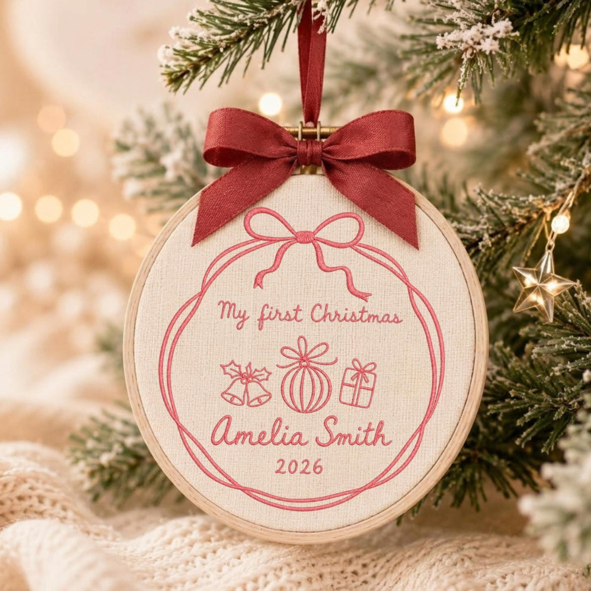May include: A round wooden embroidery hoop ornament featuring cream-colored fabric with pink embroidered text reading My first Christmas, Amelia Smith, and 2026, alongside icons of bells, a bauble, and a gift, topped with a dark red ribbon bow.