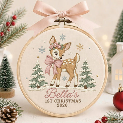 May include: A round wooden embroidery hoop containing cream fabric with a stitched design of a fawn wearing a pink bow and flower crown, flanked by two pine trees and snowflakes. The text reads Bella's 1ST CHRISTMAS 2026. A pink ribbon is tied at the top.