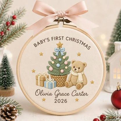 May include: A circular embroidery hoop ornament featuring a light beige fabric background with a light pink satin ribbon bow at the top. The design includes a decorated Christmas tree, a teddy bear, gift boxes, and the text Baby's First Christmas, Olivia Grace Carter, and 2026.