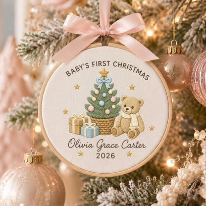 May include: A round wooden embroidery hoop ornament featuring a cream fabric background with embroidered text Baby's First Christmas, Olivia Grace Carter, and 2026. It depicts a decorated Christmas tree, a teddy bear, and gifts, topped with a pink ribbon.